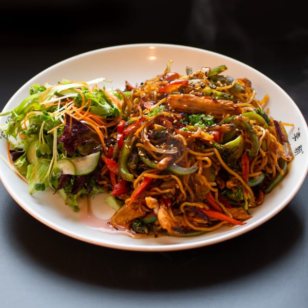 A white plate holds a serving of steaming stir-fried noodles with vegetables and a side salad.