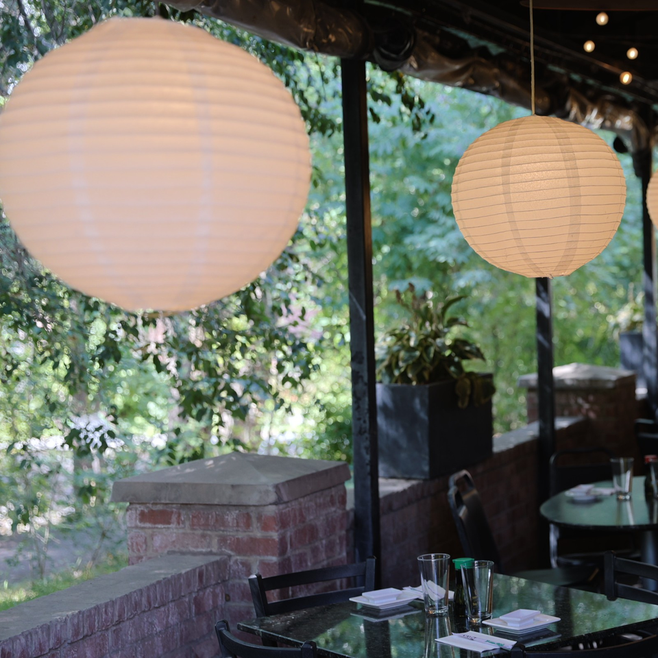 Restaurant patio with black tables, place settings, paper lanterns, brick walls, and lush greenery surrounding the dining area.